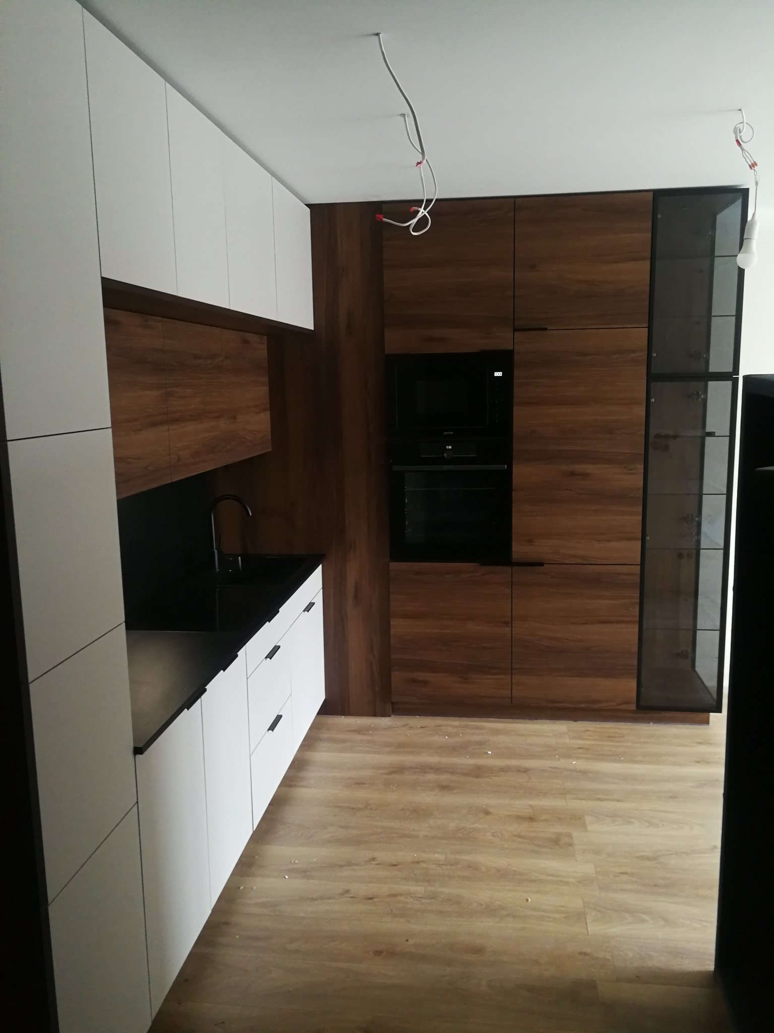 Nowoczesna kuchnia: białe szafki z czarnymi uchwytami, drewniane panele, czarny zlew i sprzęt AGD. Minimalistyczny design, surowe wykończenie, widoczne przewody.