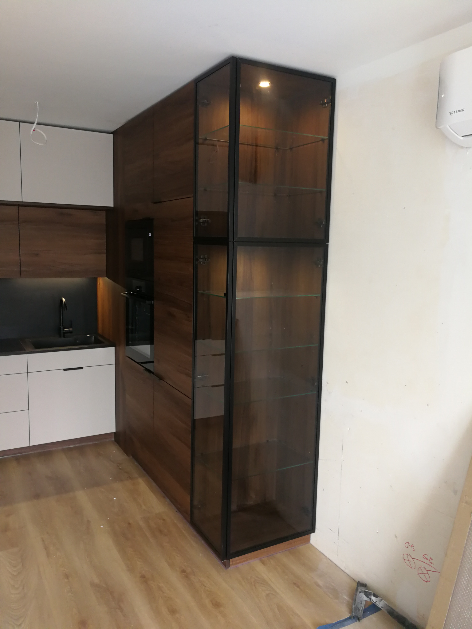 Nowoczesna kuchnia z zabudową w odcieniach bieli i drewna. Wysoka szafka z przeszklonymi drzwiami i podświetleniem. Widoczny fragment blatu i sprzęt AGD.