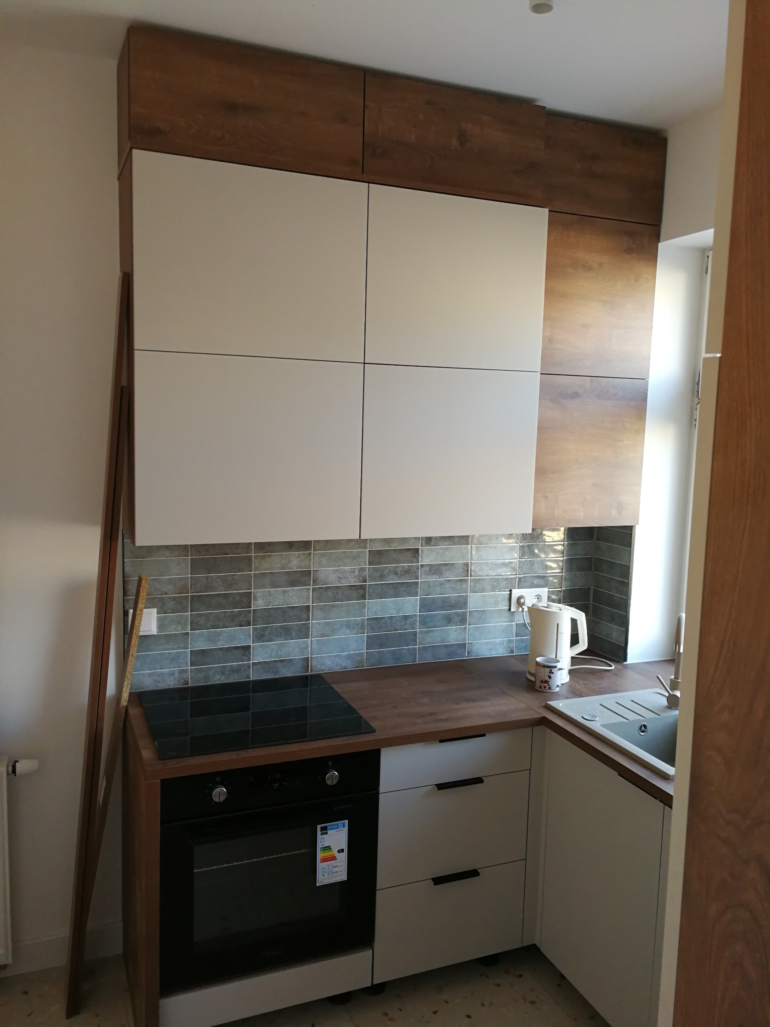 Nowoczesna kuchnia: białe szafki górne i dolne, drewniane akcenty, szare płytki nad blatem. Widoczna płyta indukcyjna i piekarnik. Minimalistyczny design.