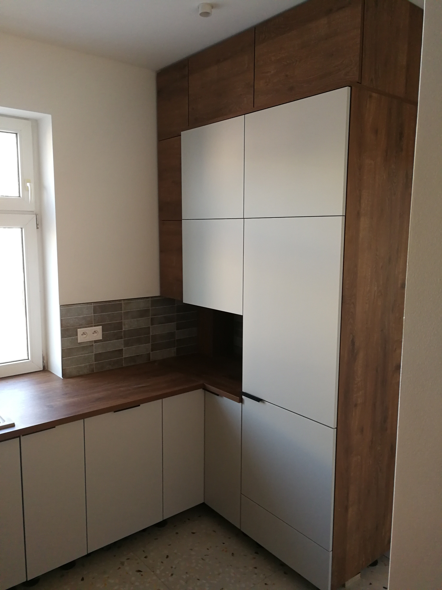 Nowoczesna kuchnia z białymi frontami szafek i drewnianym blatem, uzupełniona o brązowe szafki górne. Widoczna płytka na ścianie i fragment okna.