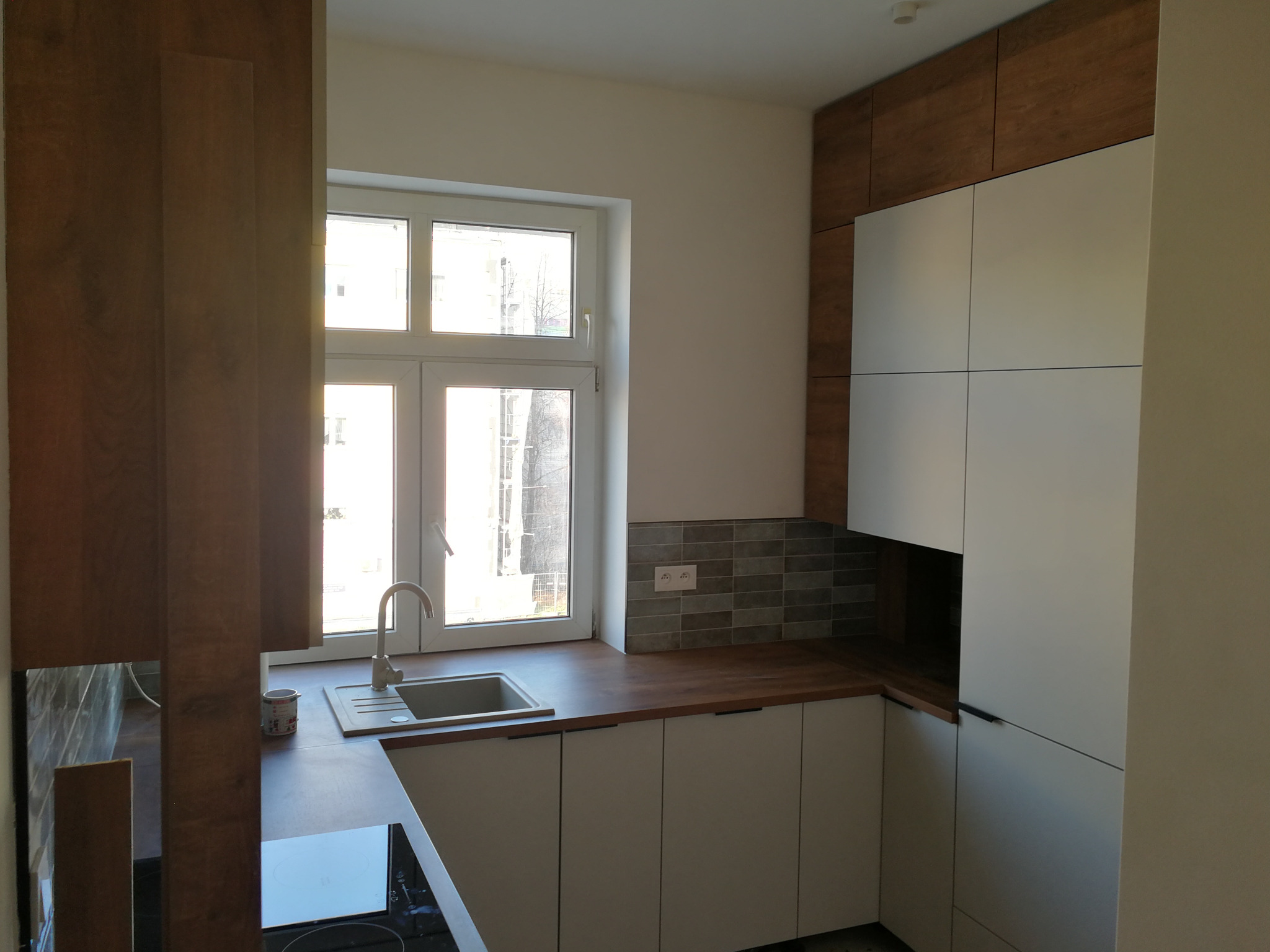 Jasna kuchnia z drewnianym blatem i białymi frontami szafek, widok na okno z widokiem na budynek. Minimalistyczny design, szare płytki nad blatem.