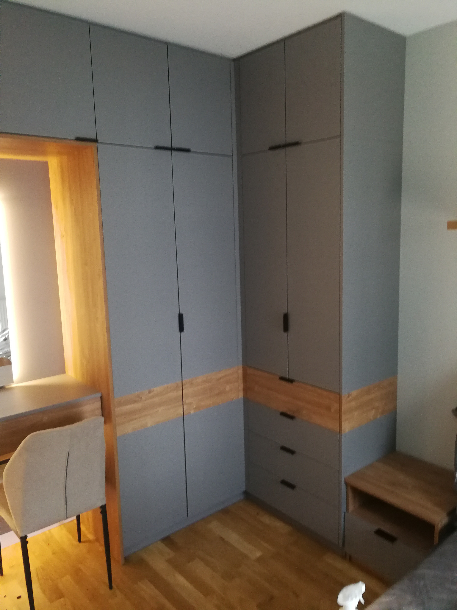 Szara szafa na wymiar z drewnianym akcentem, dopasowana do biurka i szafki nocnej, tworząca spójny design wnętrza. Minimalistyczne uchwyty i drewniana podłoga.