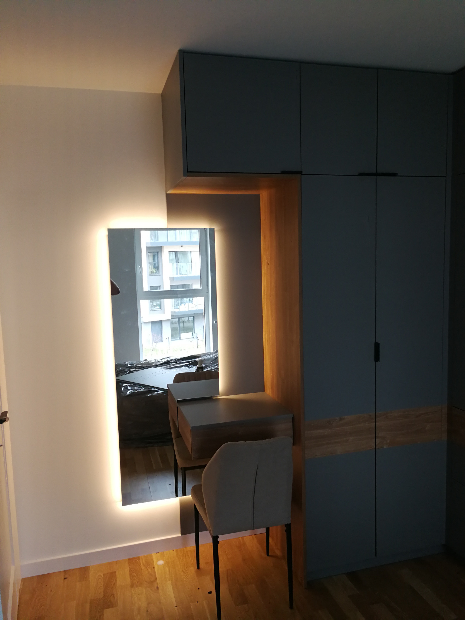 Szafa na wymiar z toaletką i lustrem z podświetleniem LED. Widoczne drewniane elementy i krzesło. W lustrze odbicie okna i fragmentu wnętrza.