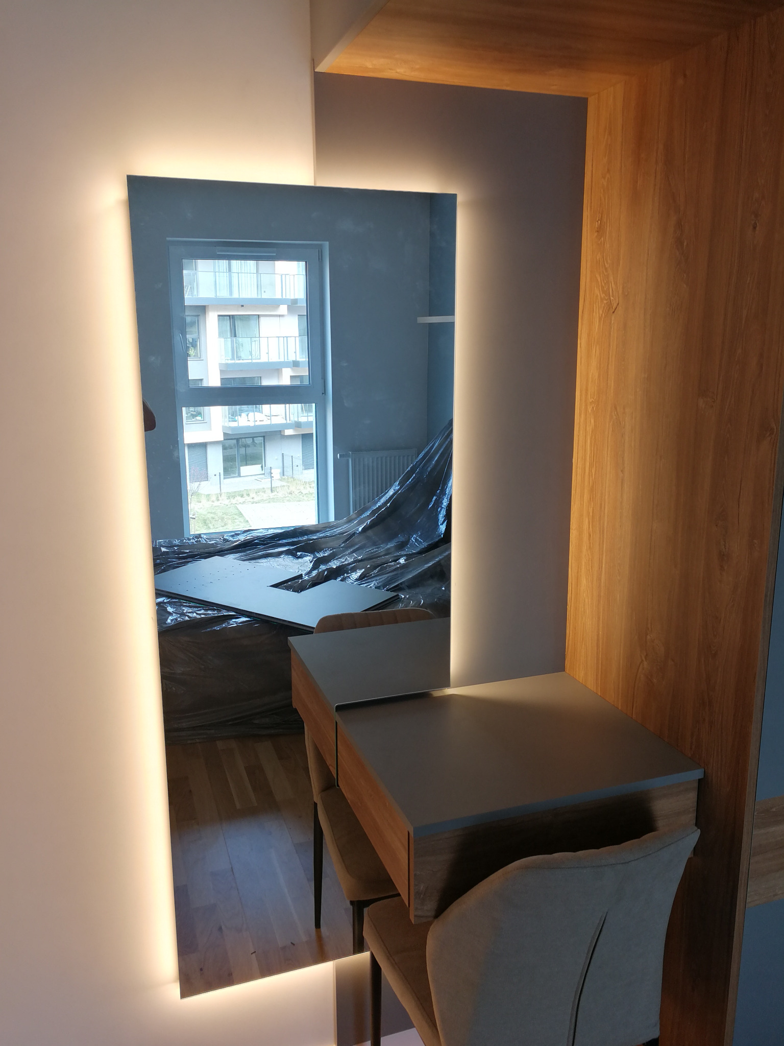 Toaletka z podświetlanym lustrem wbudowana w drewnianą zabudowę, widoczna w trakcie prac wykończeniowych. W tle okno i zabezpieczone folią meble.