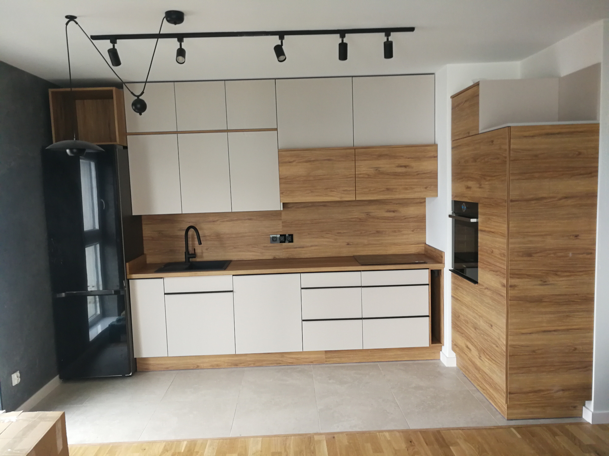 Nowoczesna kuchnia: białe szafki z drewnianymi akcentami, czarny zlew i lodówka, oświetlenie szynowe. Minimalistyczny design, jasna podłoga, mieszkanie po remoncie.