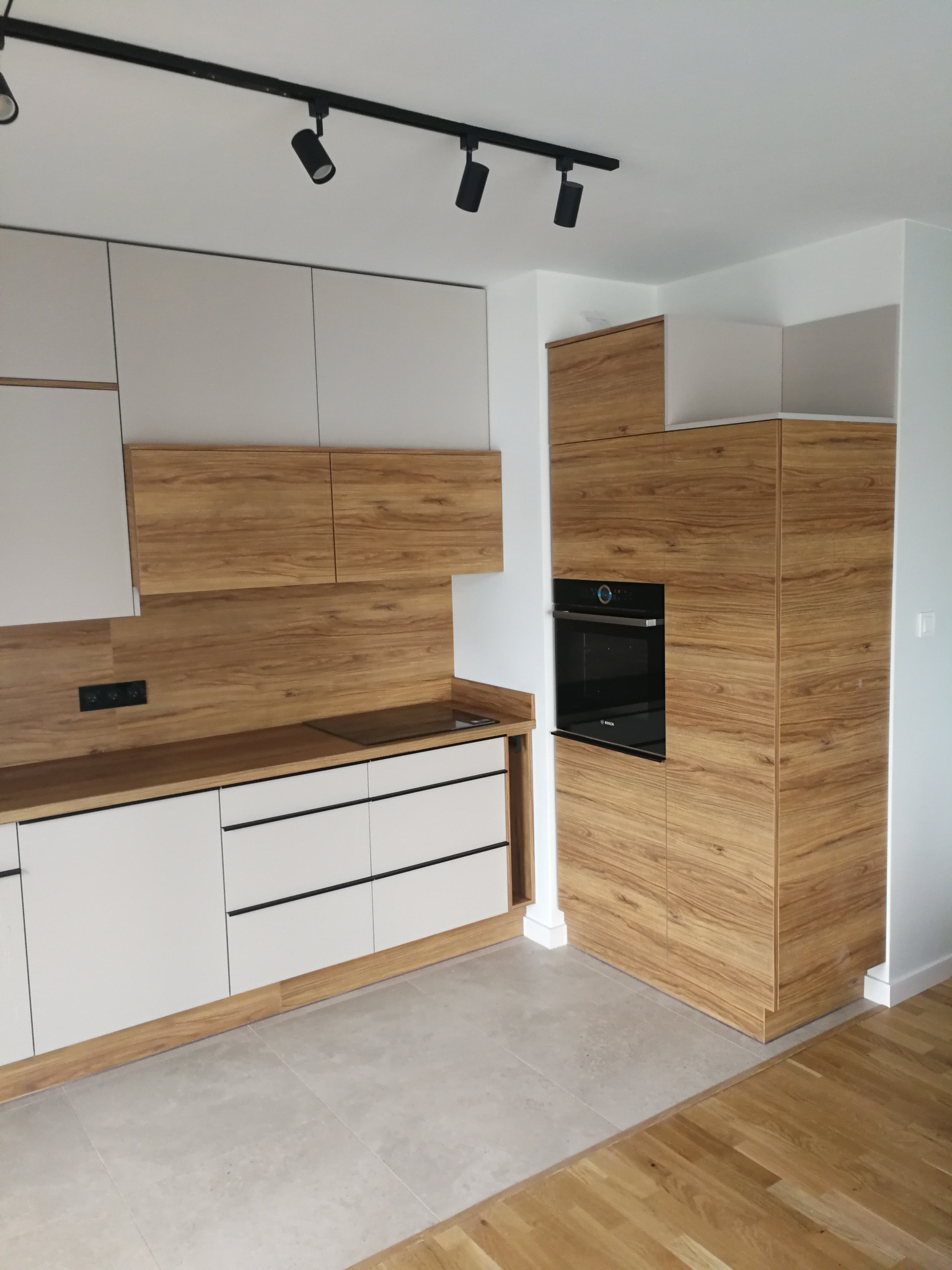 Nowoczesna kuchnia: białe szafki dolne z czarnymi uchwytami, górne szafki i zabudowa piekarnika w okleinie drewnopodobnej. Oświetlenie punktowe.