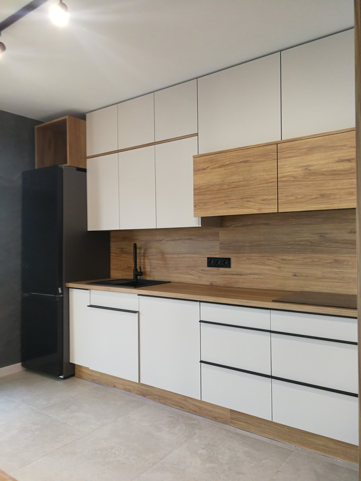 Nowoczesna kuchnia z białymi szafkami i drewnianymi akcentami. Czarna armatura i lodówka kontrastują z jasnymi frontami. Minimalistyczny design, jasna podłoga.