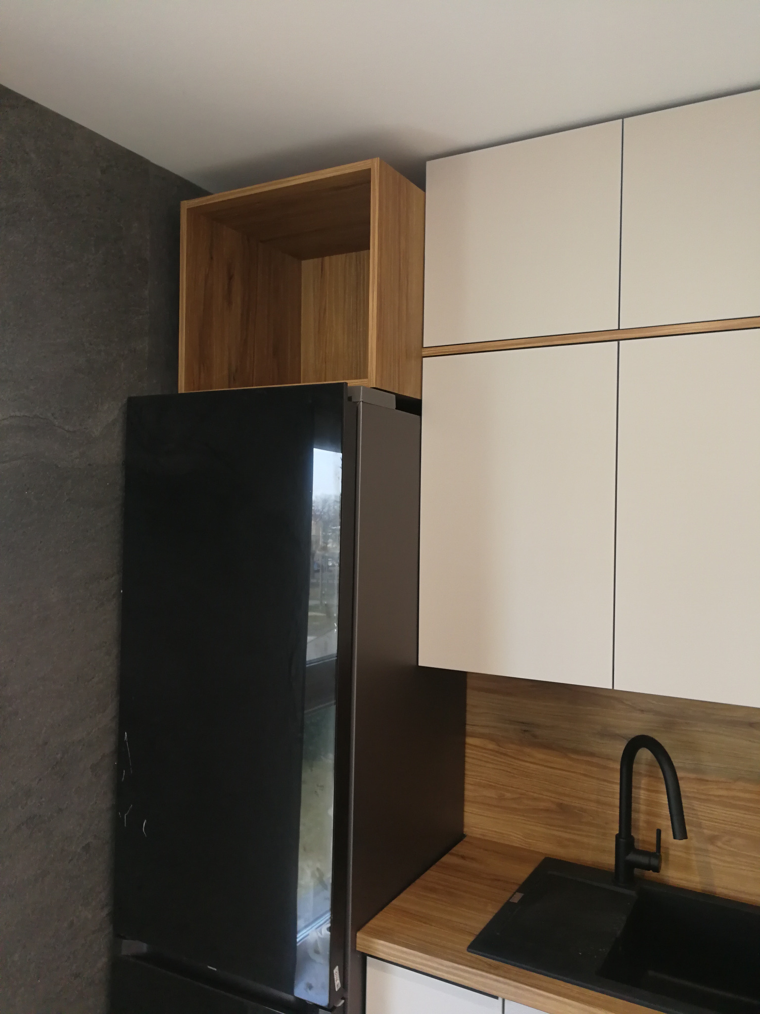 Nowoczesna kuchnia: białe szafki wiszące z drewnianymi akcentami, czarny zlew i bateria, lodówka z szafką nad nią. Kontrastowe tło z szarej ściany.