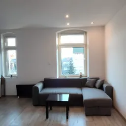 Jasne wnętrze pokoju po odnowieniu, z panelami podłogowymi imitującymi drewno, szary narożnik i czarny stolik kawowy, widoczne okna z białymi ramami i punktowe oświetlenie sufitowe.