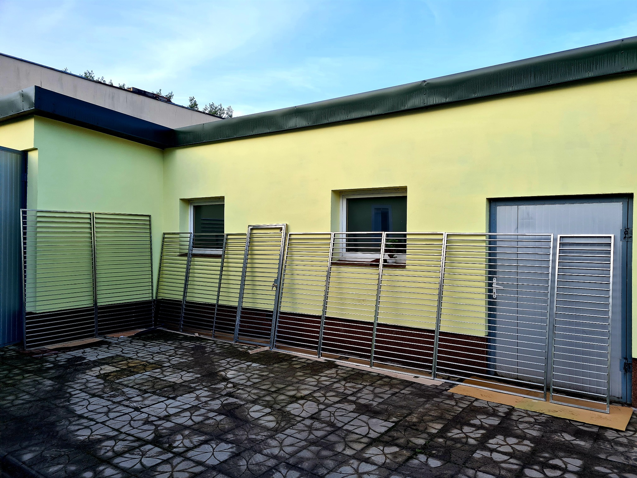 Przygotowane do montażu, surowe, metalowe segmenty ogrodzeniowe o poziomym układzie szczebli, oparte o ścianę budynku z żółtą elewacją i szarą stolarką okienną i drzwiową, na nierównej, mokrej...