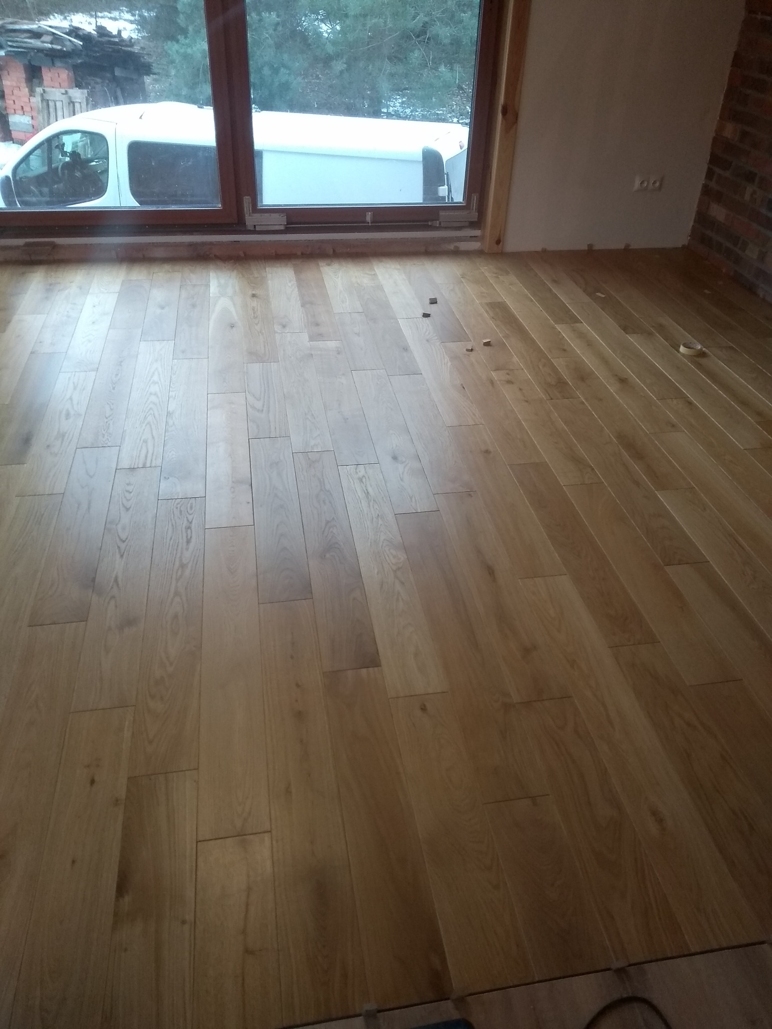 Jasne, drewniane deski podłogowe ułożone w jodełkę, widoczne kołki dystansowe i taśma malarska, okno z widokiem na biały samochód dostawczy w zimowym krajobrazie, surowy mur z cegły w tle.