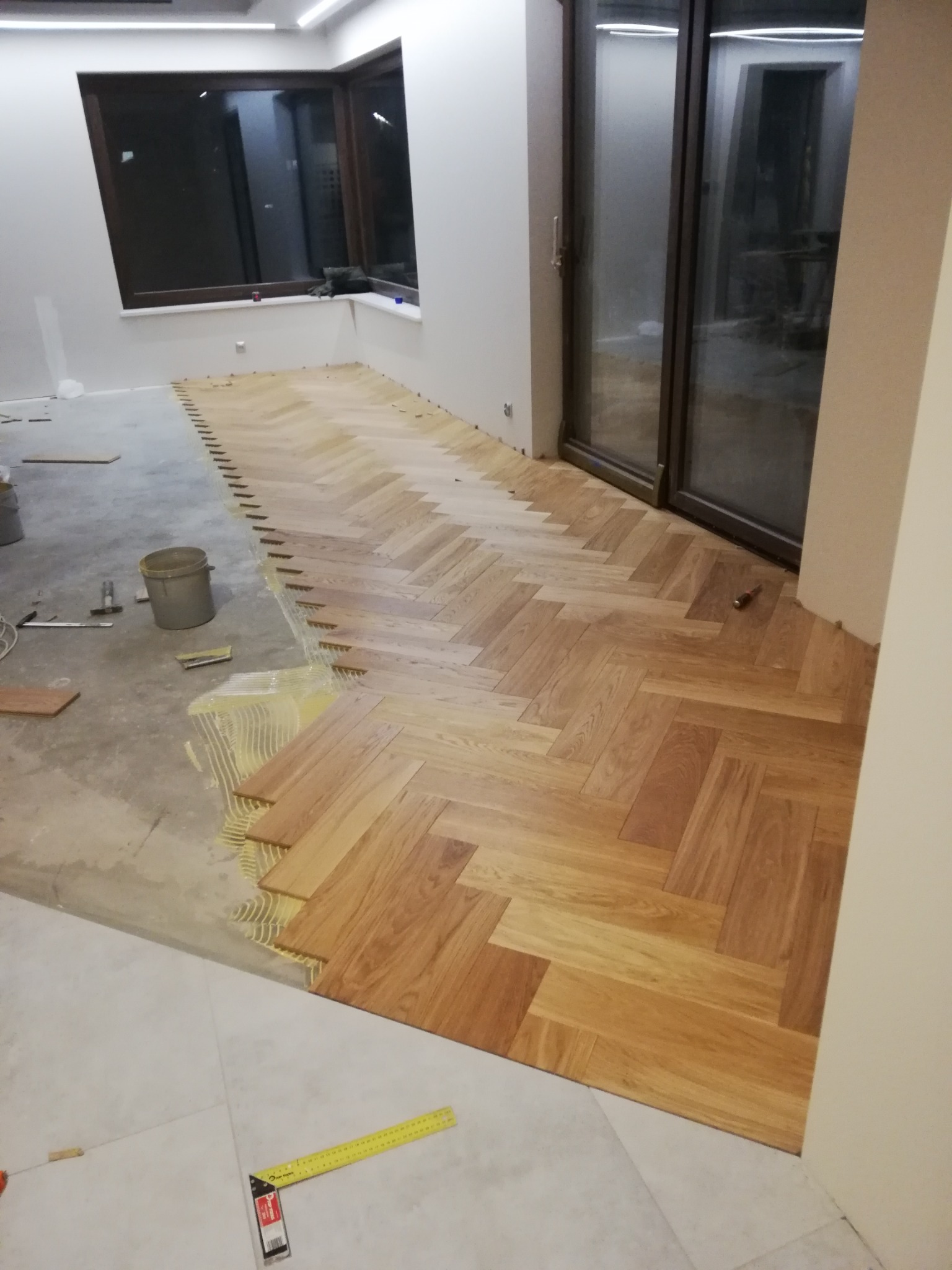 Fragment podłogi w trakcie układania parkietu w jodełkę, widoczne klej, narzędzia i surowy beton obok ułożonej części, okno i drzwi balkonowe w tle.