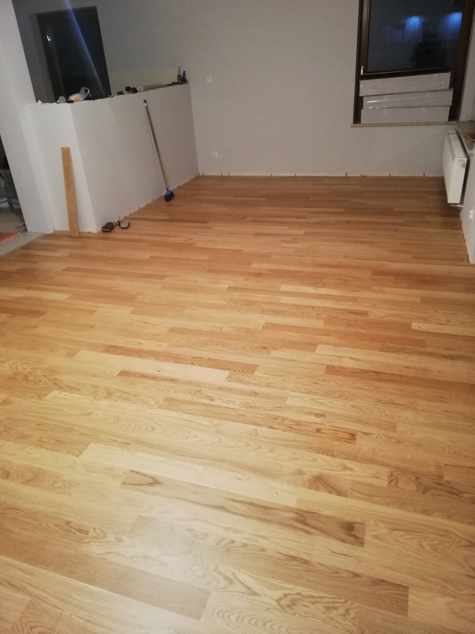 Jasny, drewniany parkiet ułożony w nowym pomieszczeniu, widoczne listwy przypodłogowe oraz okno z zapakowanymi roletami.