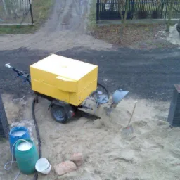 Żółta maszyna do wylewania jastrychu na przyczepce, z widocznymi dwoma beczkami z płynem, łopatą i stertą piasku na placu budowy.