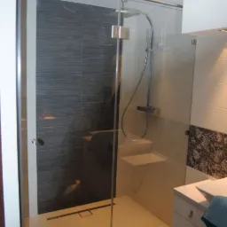 Nowoczesna kabina prysznicowa z transparentnego szkła, chromowane okucia, szara ściana z poziomymi paskami, mała półka wewnątrz kabiny, odpływ liniowy w podłodze.