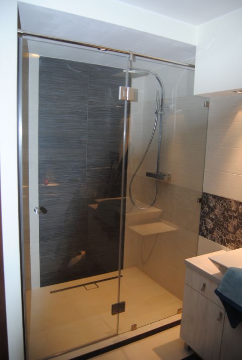 Nowoczesna kabina prysznicowa z transparentnego szkła, chromowane okucia, szara ściana z poziomymi paskami, mała półka wewnątrz kabiny, odpływ liniowy w podłodze.