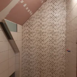 Ściana łazienki w trakcie wykańczania z ułożonymi płytkami o wzorze geometrycznym, widoczne fragmenty sufitu ze śladami gładzi oraz obudowa pionu z płyt gipsowo-kartonowych.