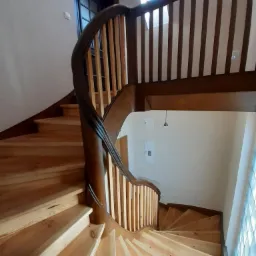 Drewniane, spiralne schody z ciemnym poręczem i jasnymi stopniami, widok z góry, okno dachowe w tle.