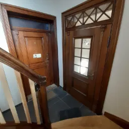 Klatka schodowa z drewnianymi schodami i dwoma drewnianymi drzwiami wejściowymi do mieszkań, z których jedne mają numer 1 i małą tabliczkę z psem rasy Husky.