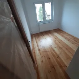 Jasne, drewniane panele podłogowe w pokoju z białymi ścianami i oknem; widoczna folia ochronna na drzwiach.