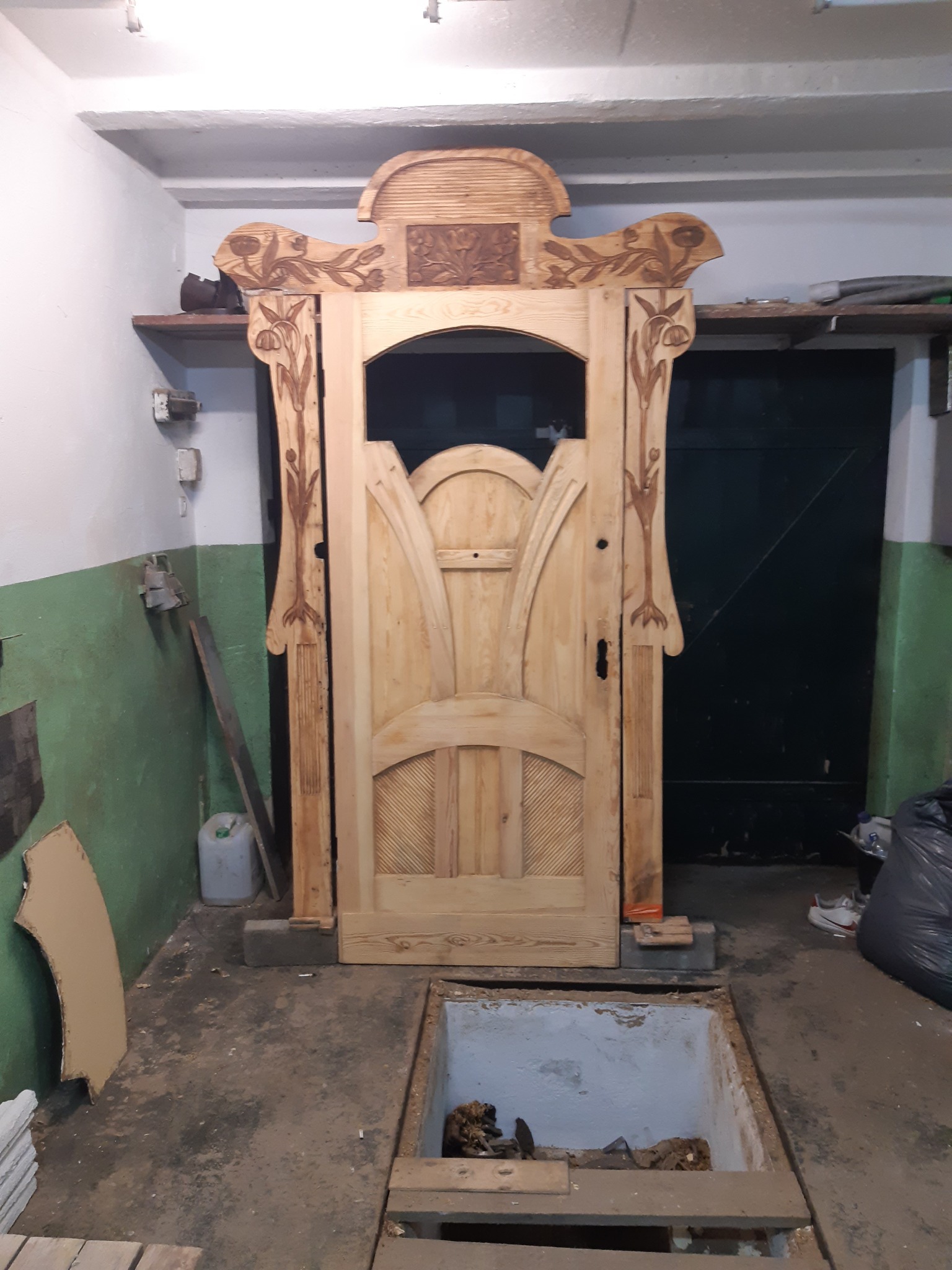 Surowa, rzeźbiona, drewniana rama drzwiowa z motywami roślinnymi, ustawiona w surowym wnętrzu z odkrytą studzienką podłogową.