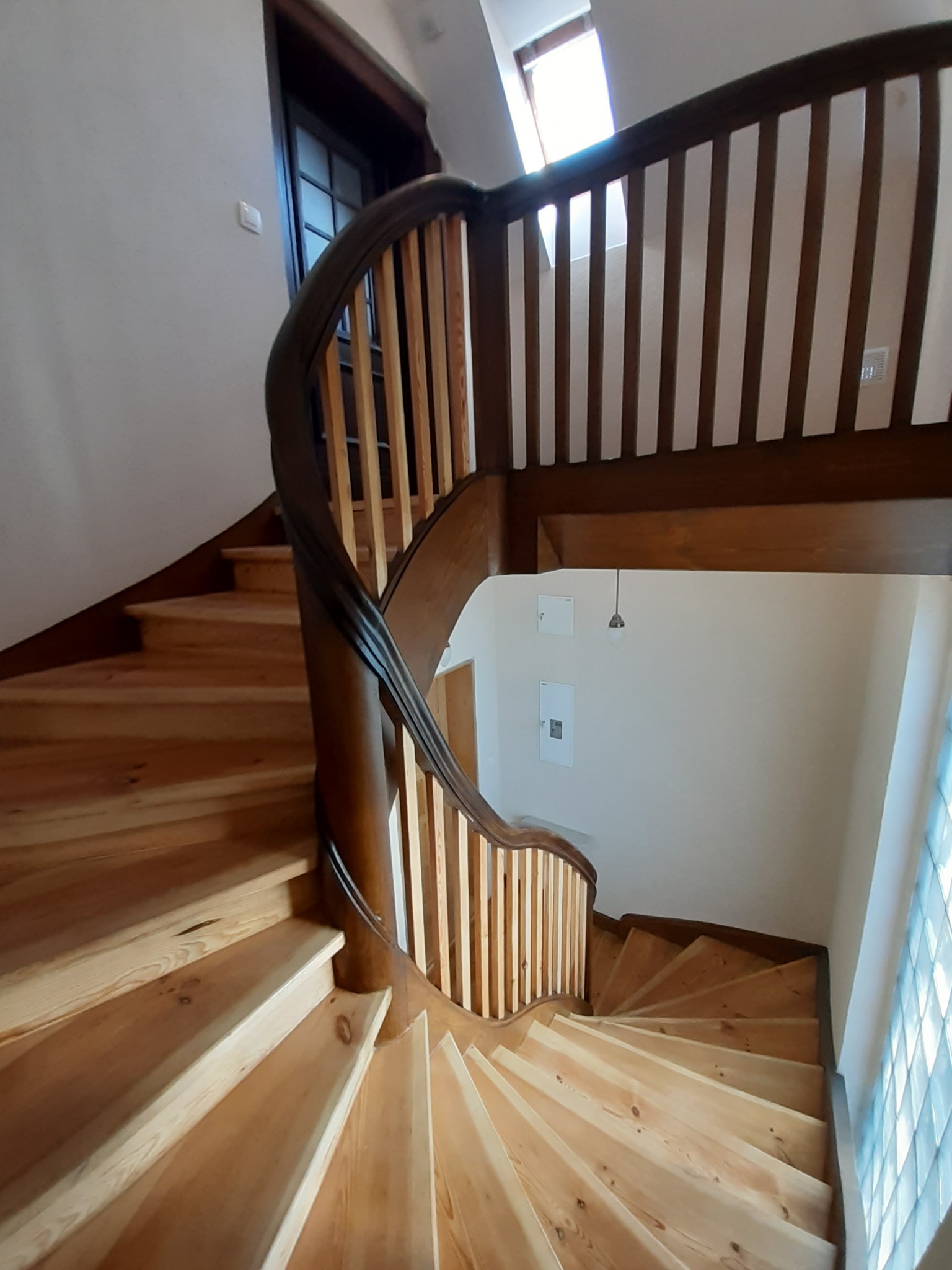 Drewniane, spiralne schody z ciemnym poręczem i jasnymi stopniami, widok z góry, okno dachowe w tle.