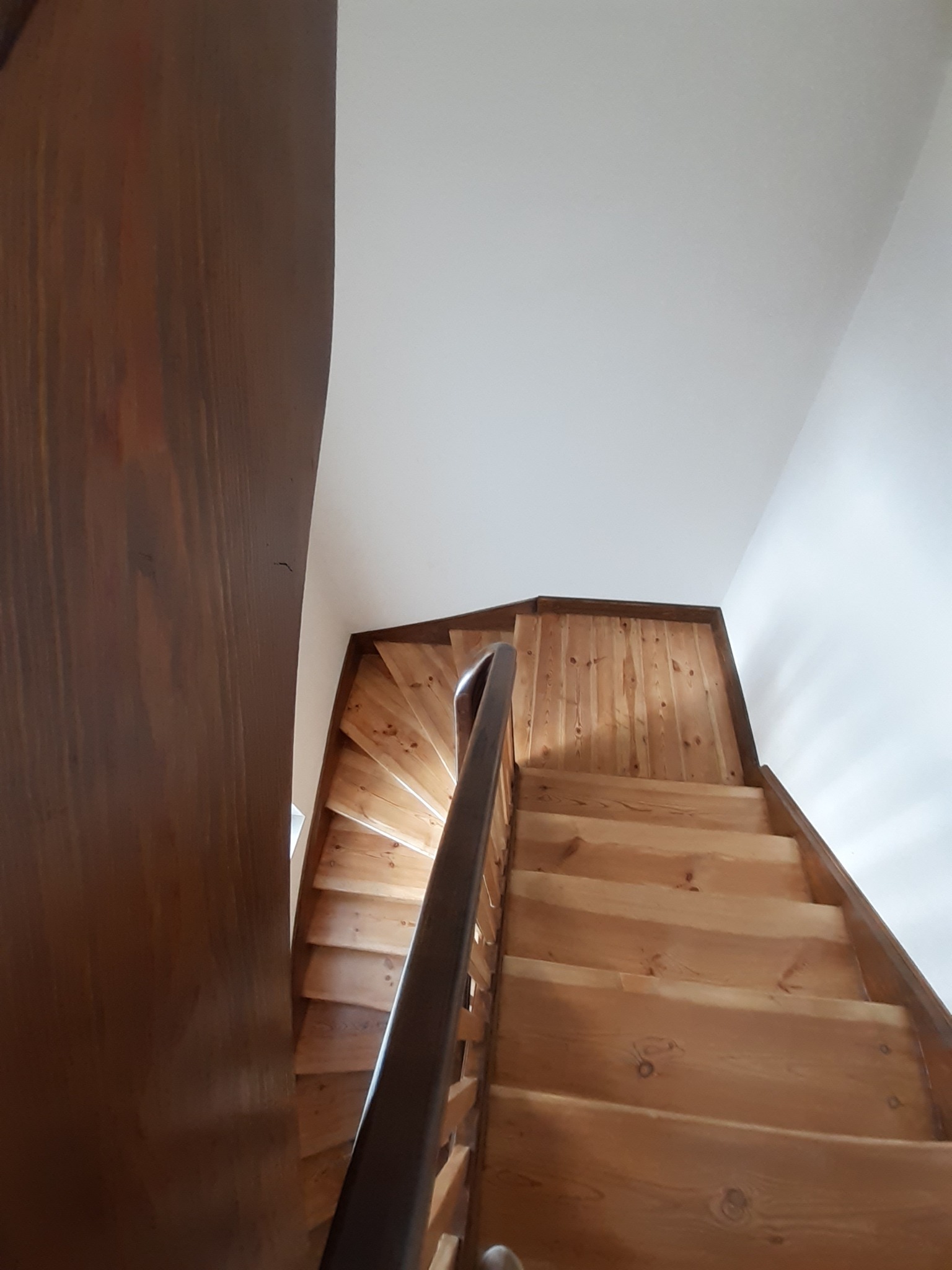 Drewniane schody spiralne z ciemną balustradą, widok z dołu, z fragmentem ciemnego futryny drzwi po lewej stronie.