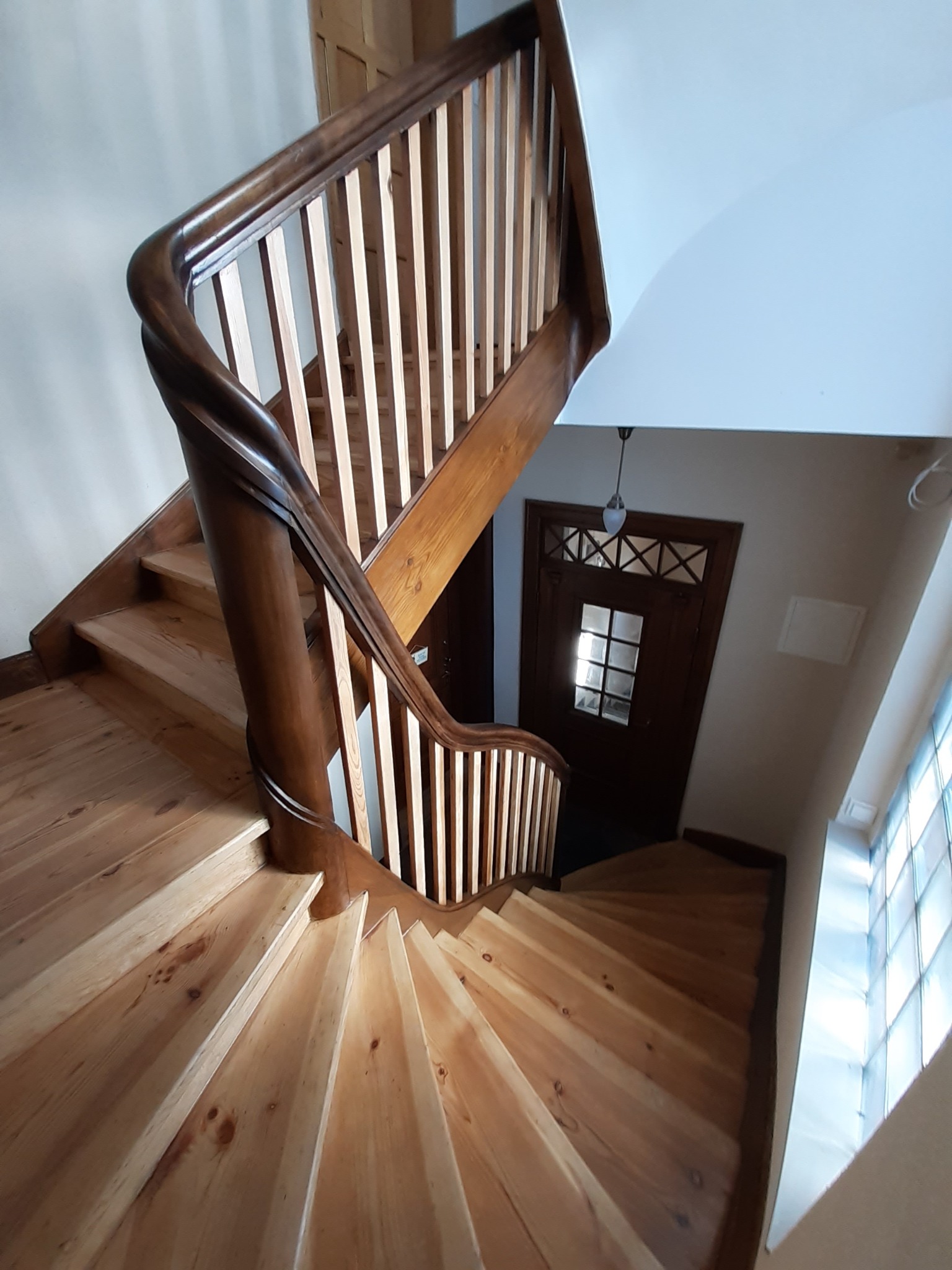 Drewniane schody spiralne z ciemnobrązową balustradą, widok z góry, z widocznym fragmentem drzwi wejściowych i okna z luksferami.
