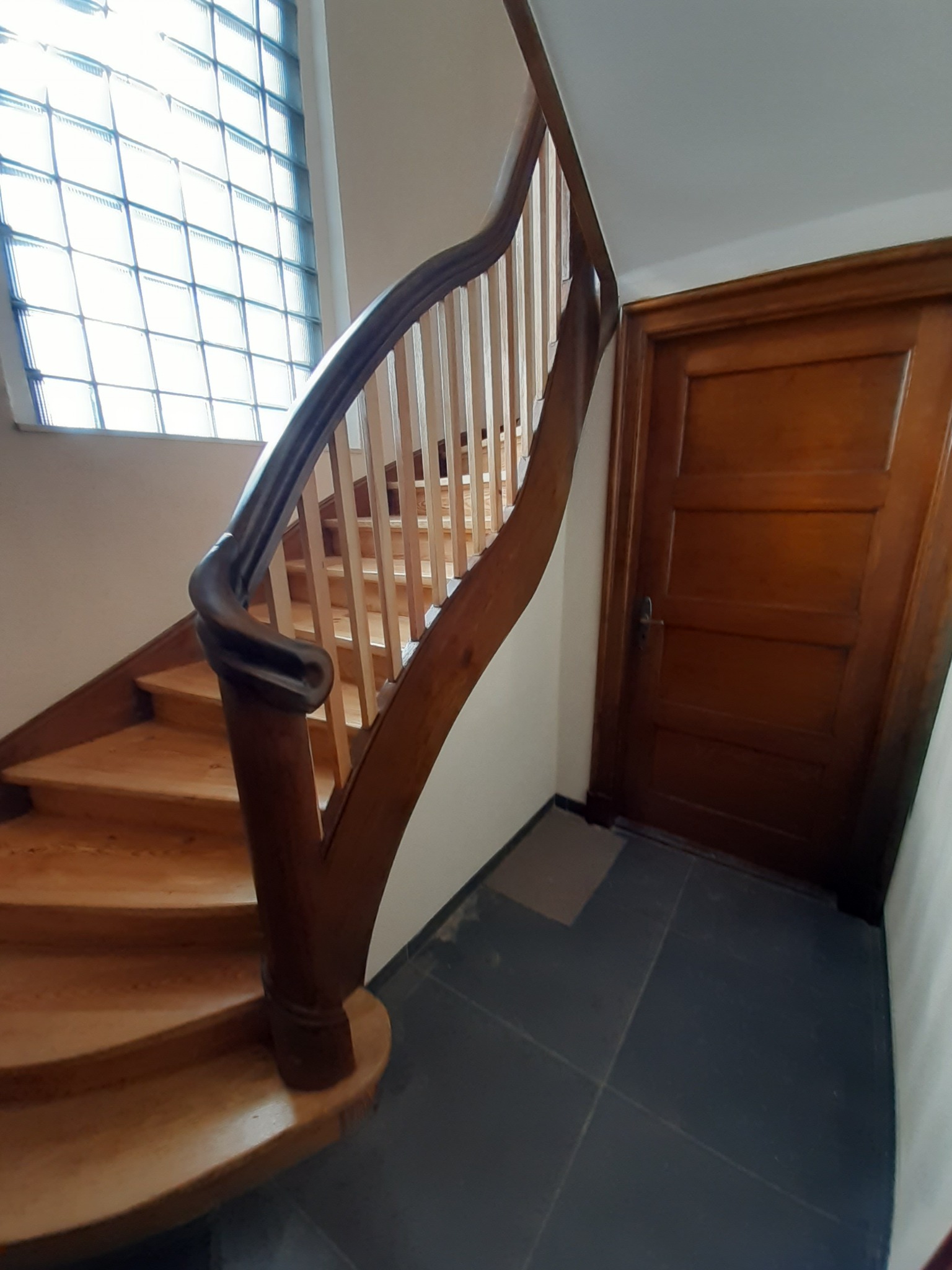 Klatka schodowa z drewnianymi schodami i balustradą, widoczna brązowa drewniana futryna drzwi, okno z luksferami i ciemna podłoga.