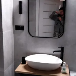 Nowoczesna łazienka z owalną umywalką nablatową, czarną armaturą i lustrem w czarnej ramie. Na blacie dozownik do mydła 'Biały Jeleń'. W lustrze odbija się osoba robiąca zdjęcie telefonem.