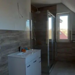 Ukończona łazienka na poddaszu z prysznicem typu walk-in, umywalką z szafką i grzejnikiem drabinkowym, wykończona płytkami imitującymi drewno i białymi płytkami, widoczne wystające kable elektryczne.