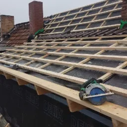 Konstrukcja drewniana dachu w trakcie budowy, widoczna membrana Delta Vent Neo, fragment dachu pokryty starą dachówką, kominy z cegły, na belce spoczywa piła tarczowa Hikoki.