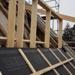 Konstrukcja drewniana dachu w trakcie wymiany, widoczna membrana dachowa Delta i narzędzie do montażu, w tle rusztowanie i drzewa.