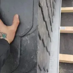 Układanie łupka na krawędzi dachu, widoczna dłoń z tatuażem i fragment konstrukcji drewnianej.