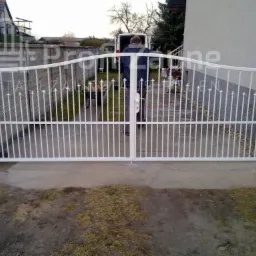 Biała, dwuskrzydłowa brama wjazdowa z metalowymi prętami i łukowatym górnym profilem, widok z zewnątrz na podwórze z zaparkowanym busem.