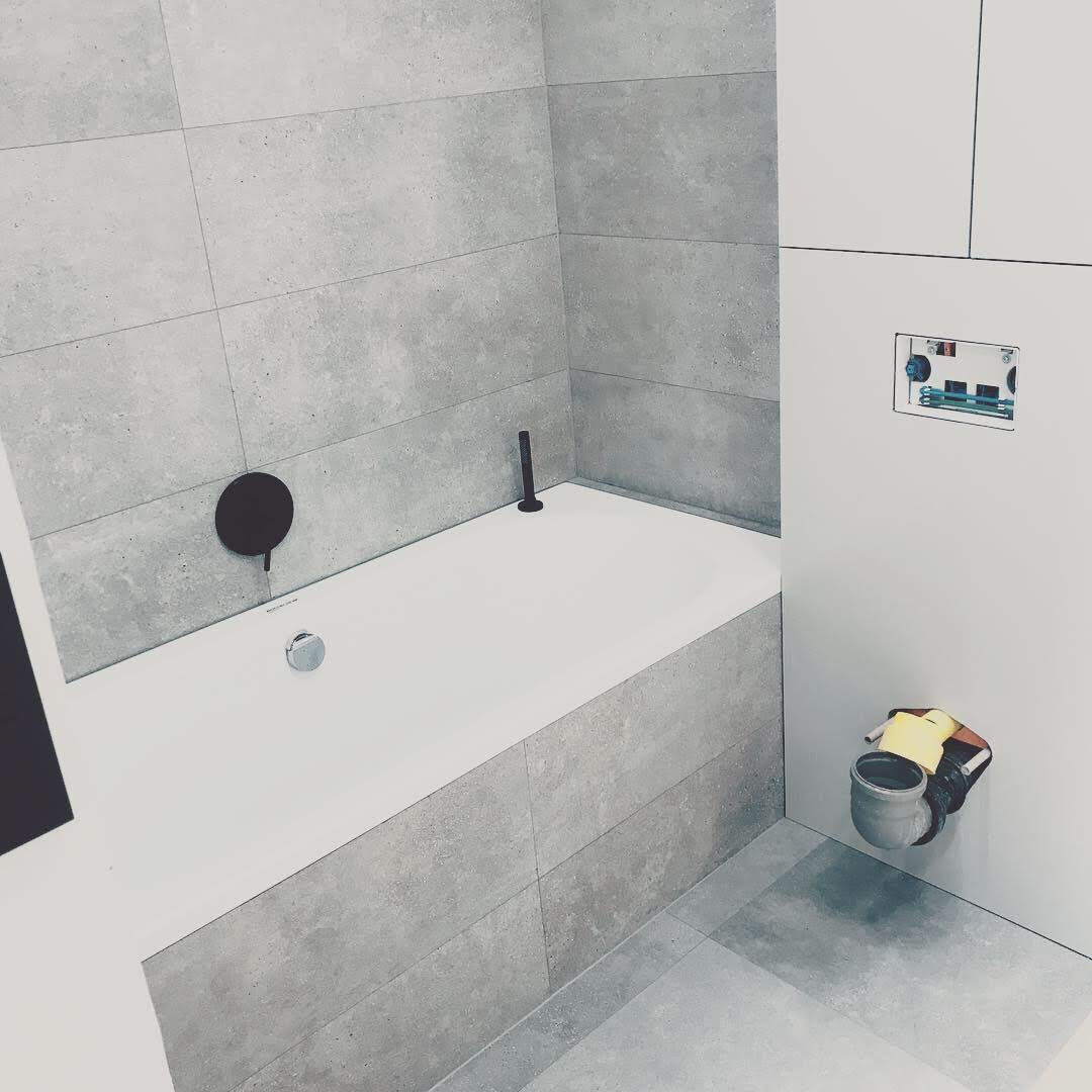 Nowoczesna łazienka w szarościach z białą wanną obudowaną płytkami, czarna armatura, widoczny etap prac instalacyjnych z wiadrem i narzędziami.