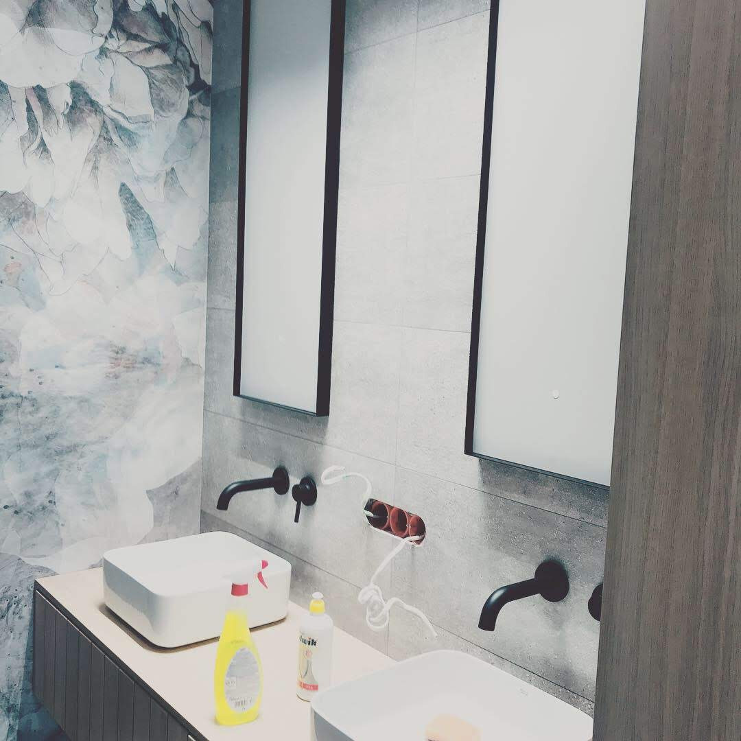 Łazienka z dwoma umywalkami nablatowymi, czarną armaturą, dużymi lustrami w ciemnych ramach i dekoracyjną tapetą w kwiaty na jednej ścianie. Widoczne niedokończone podłączenie elektryczne.