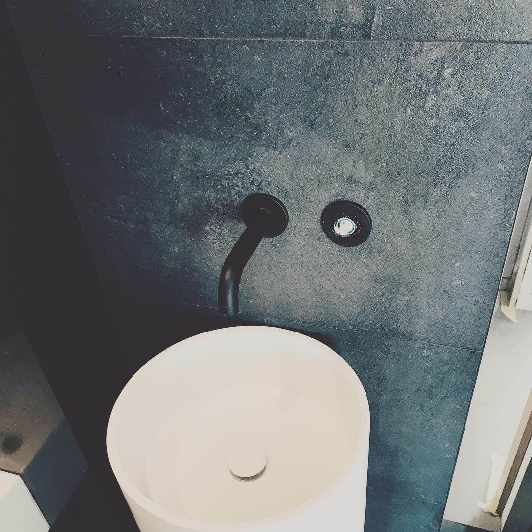 Biała, okrągła umywalka nablatowa z czarną, matową armaturą na tle ciemnoszarej, kamiennej ściany w nowoczesnej łazience.