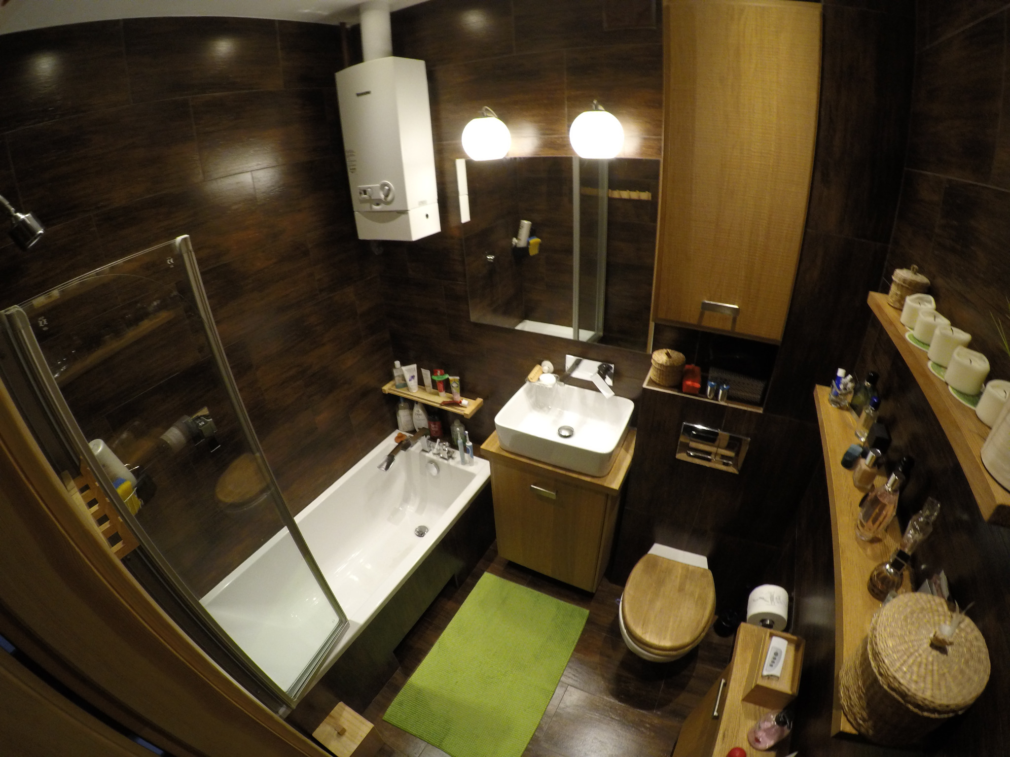 Ciemna łazienka z brązowymi płytkami, białą wanną z parawanem, umywalką na drewnianej szafce, drewnianą toaletą, podgrzewaczem wody i akcesoriami na półkach.