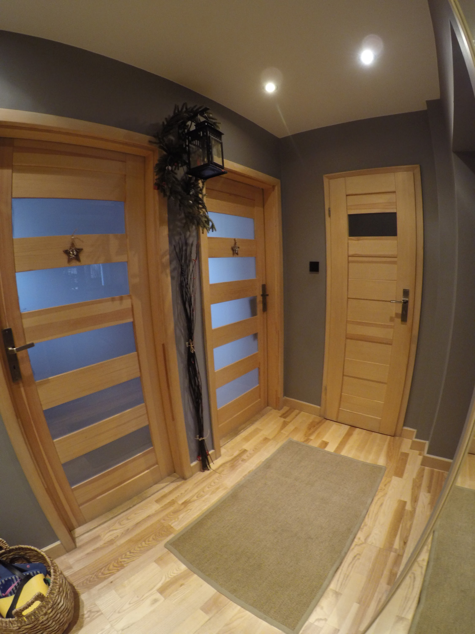 Wnętrze przedpokoju z trzema drewnianymi drzwiami, dwiema z przeszkleniami i dekoracyjnymi gwiazdkami, oraz lampionem z zielenią, na podłodze jasny parkiet i dywanik.