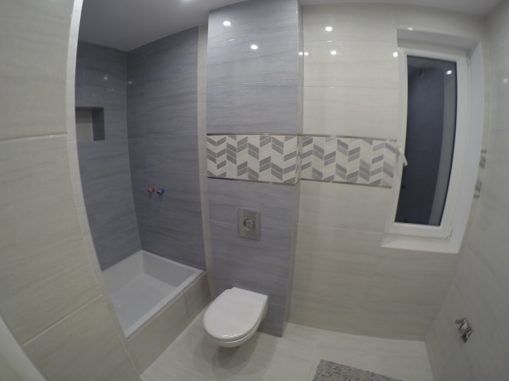 Wykończona łazienka z szarymi i białymi płytkami, wzorzysty pas dekoracyjny, biała armatura i okno.