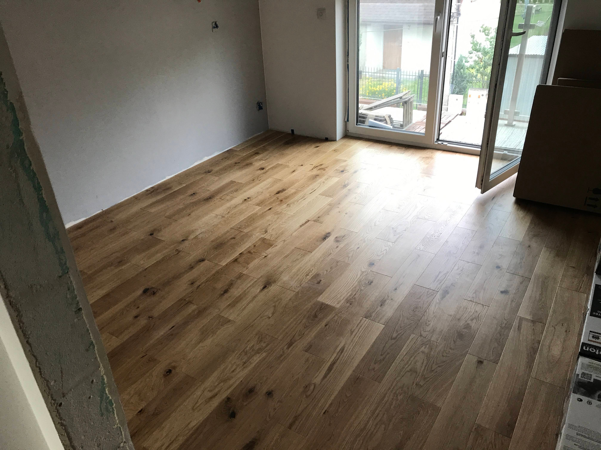 Ułożona drewniana podłoga w jasnym odcieniu dębu w pokoju z przygotowanymi ścianami do dalszych prac wykończeniowych, widoczne wystające kable elektryczne i otwarte drzwi balkonowe.