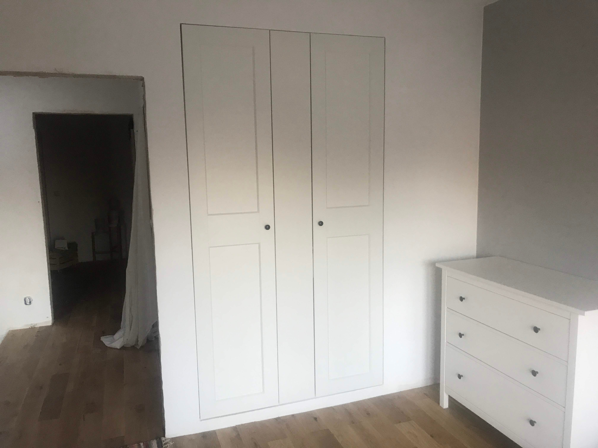 Biała, dwudrzwiowa szafa wnękowa z zamontowanymi drzwiami, widoczna w trakcie prac wykończeniowych pomieszczenia z drewnianą podłogą i białą komodą z szufladami.