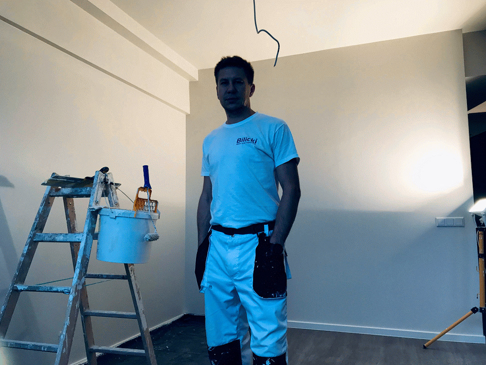 Malarz w białym stroju roboczym stoi w pokoju z pomalowanymi ścianami, drabina z wiadrem i wałkami malarskimi obok.