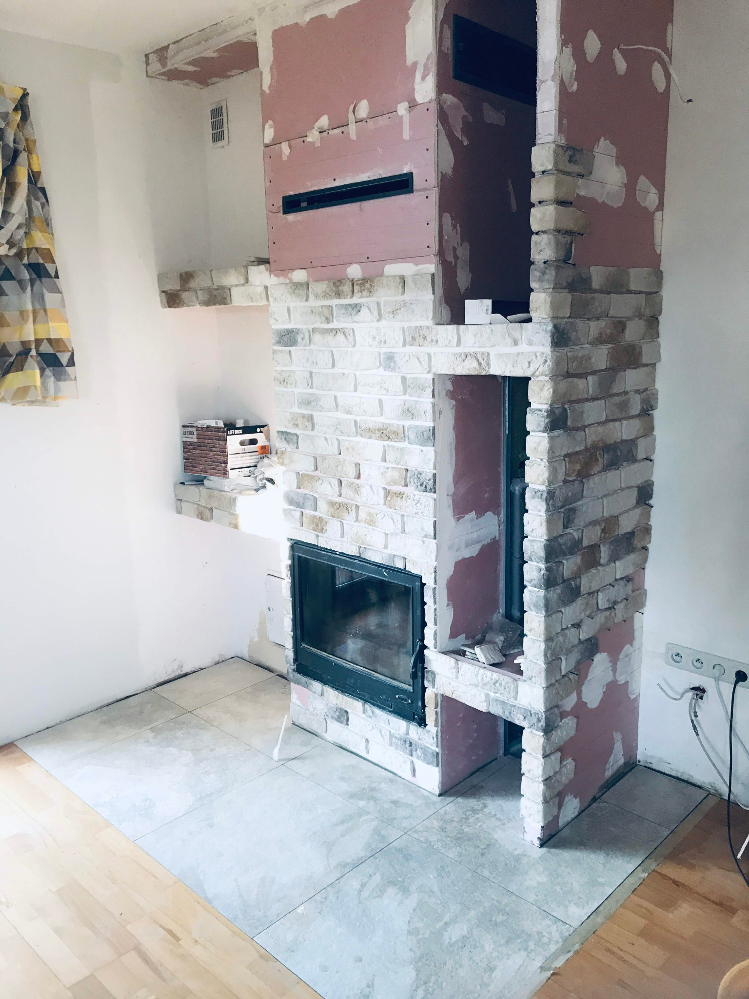 Wnętrze w trakcie remontu, z widocznym kominkiem obłożonym cegłą dekoracyjną w odcieniach bieli i szarości, ściany częściowo przygotowane do szpachlowania, podłoga wyłożona płytkami ceramicznymi...