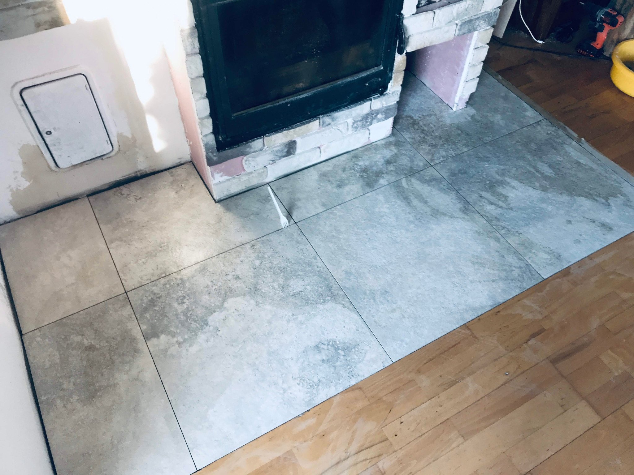 Kafelki podłogowe o szarym wzorze marmuru, ułożone wokół kominka z cegieł, z widocznym fragmentem drewnianej podłogi i narzędzia do remontu w tle.