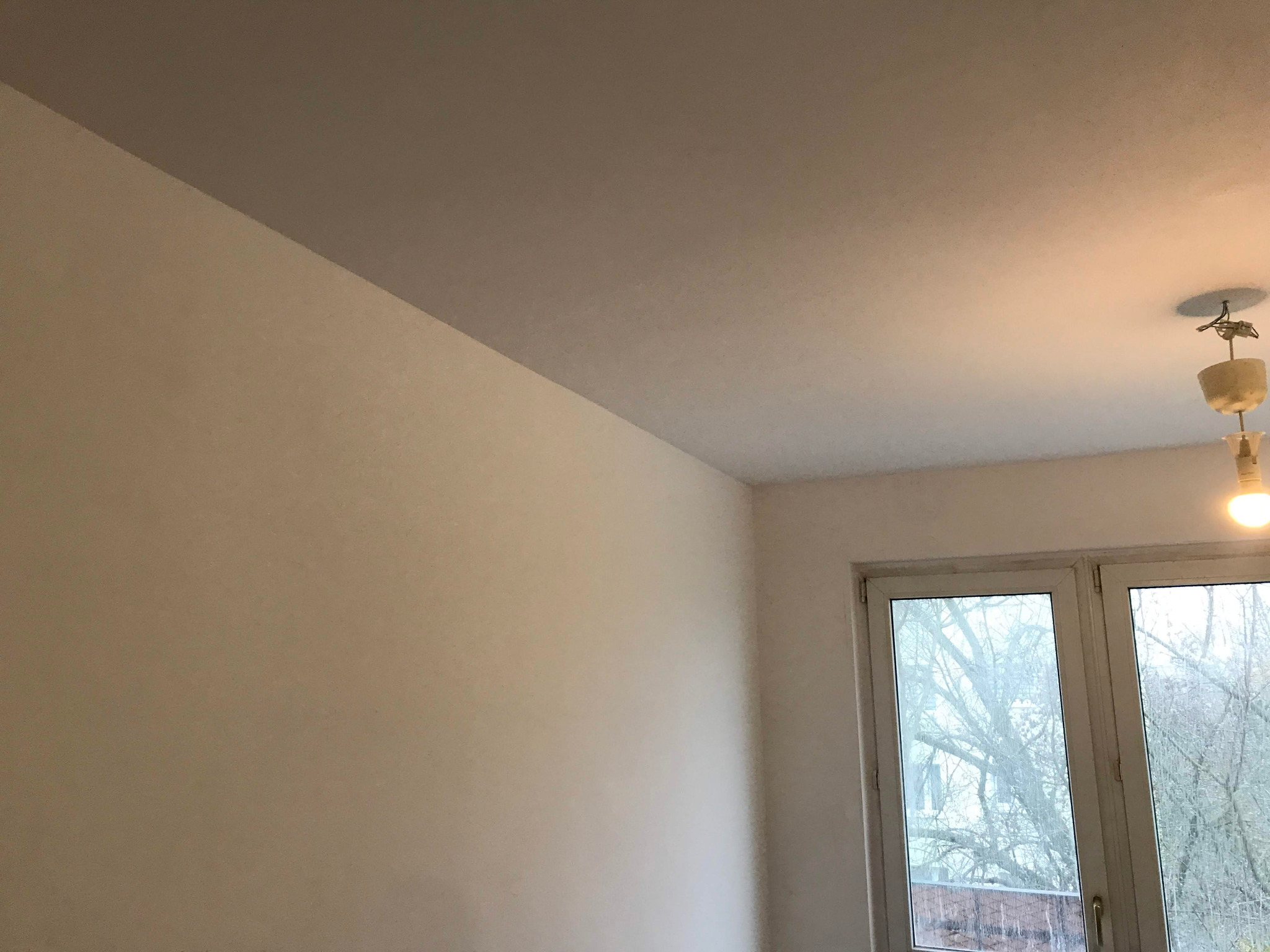 Pomieszczenie po malowaniu, widoczna ściana i sufit w dwóch odcieniach beżu, lampa sufitowa i okno z widokiem na drzewa bez liści.