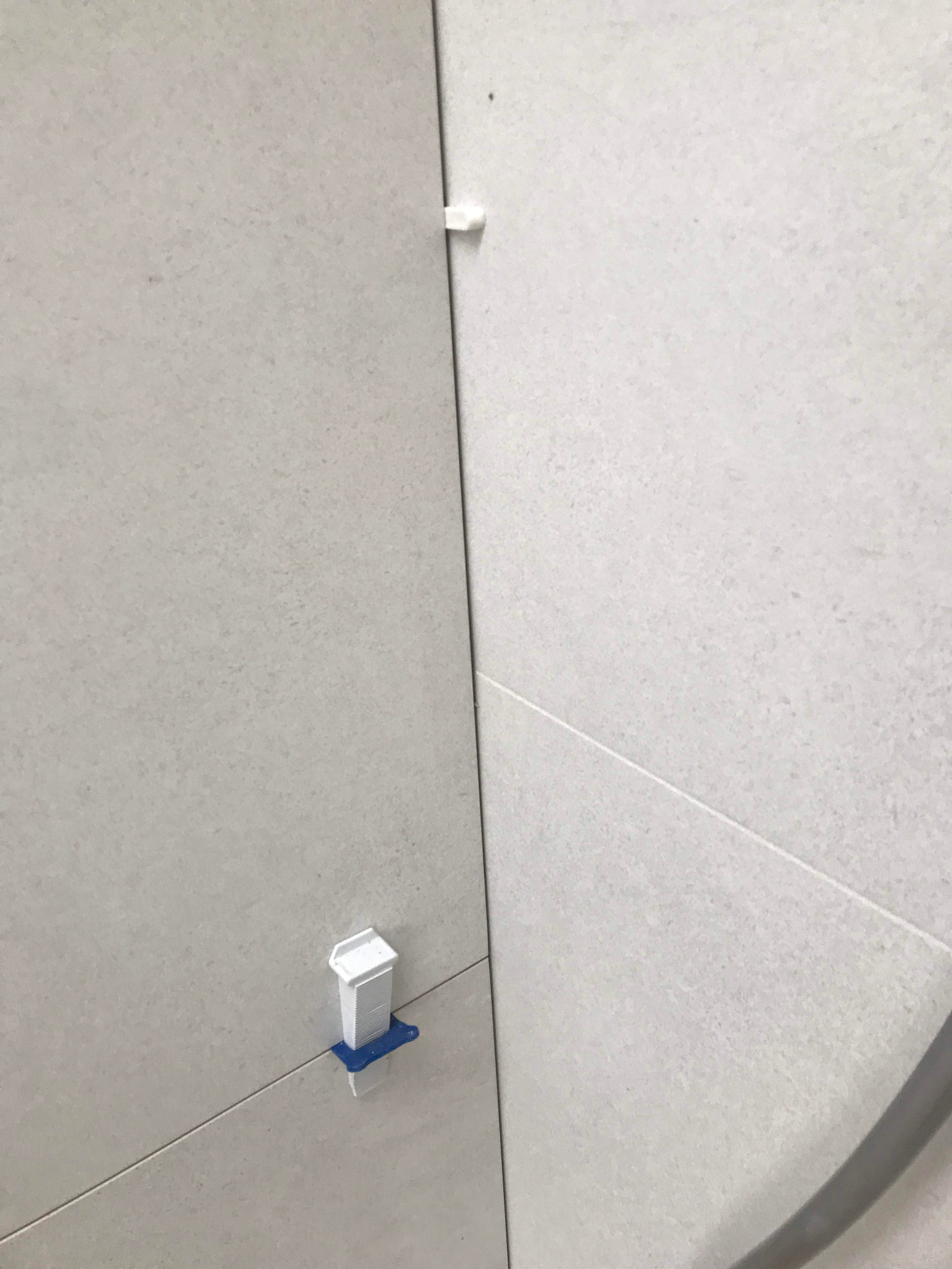 Jasne, matowe płytki ścienne ułożone z użyciem systemu poziomowania z białymi klinami i niebieskimi podstawkami, widoczne fugi.