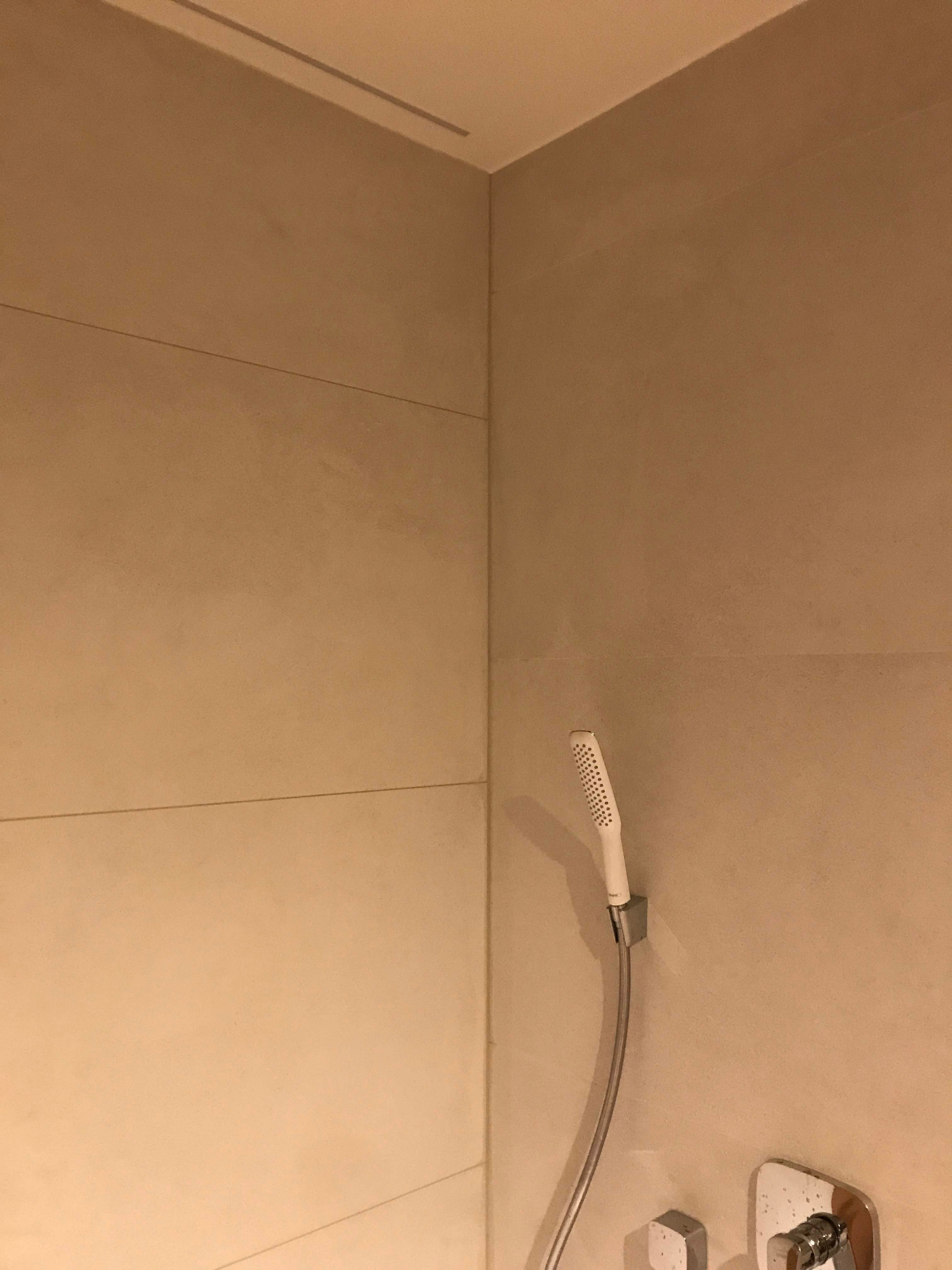 Beżowe, poziome płytki w kabinie prysznicowej z zamontowaną białą słuchawką prysznicową i chromowaną armaturą.
