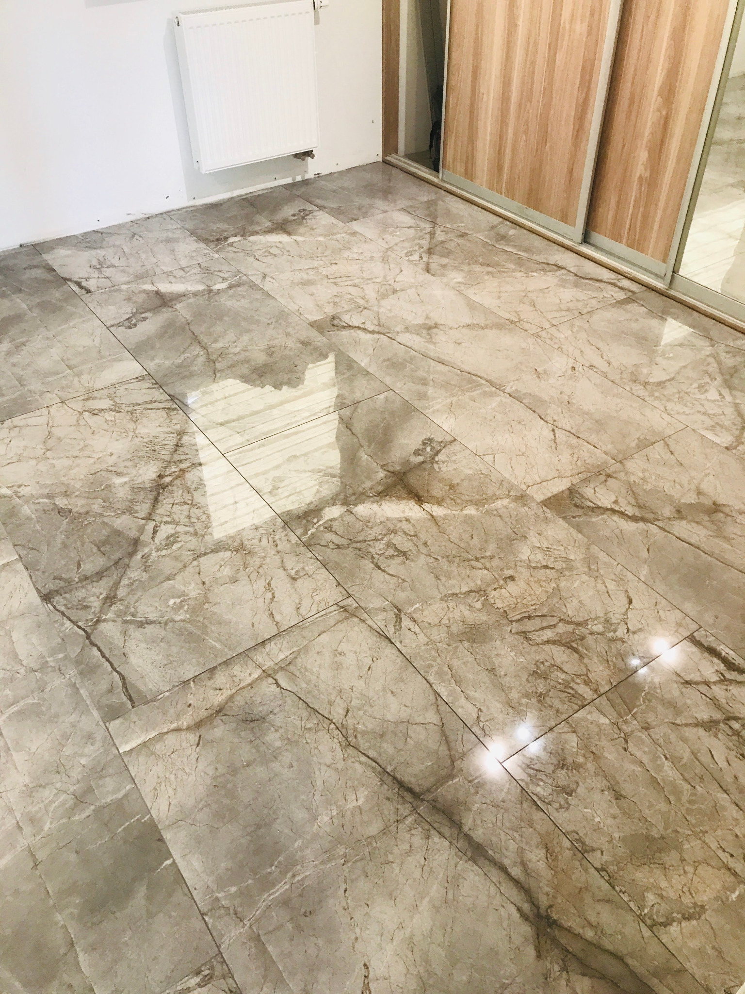 Ułożona podłoga z płytek imitujących marmur w odcieniach beżu i szarości, widoczny biały grzejnik na ścianie i fragment szafy przesuwnej z drewnianymi drzwiami.